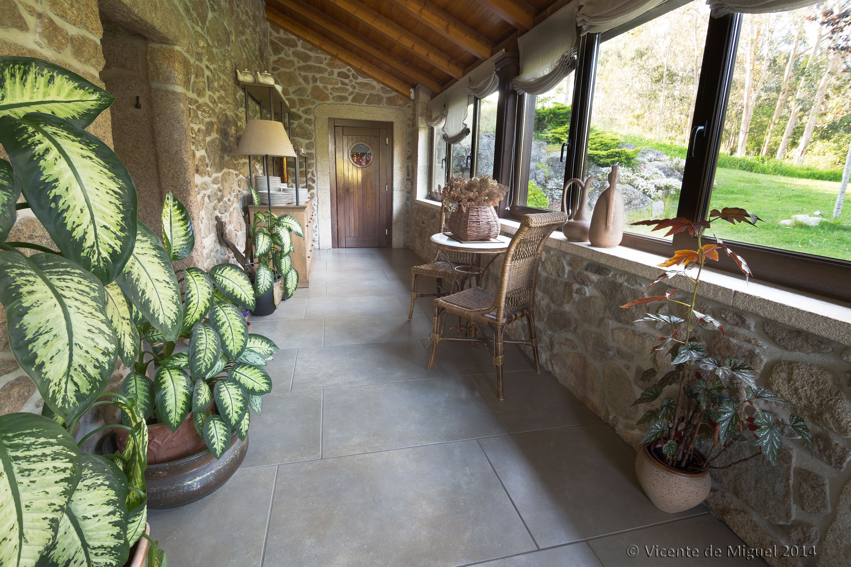 Un couloir aux murs de pierre, avec un plafond en bois, une chaise en osier, une table et des plantes en pot.