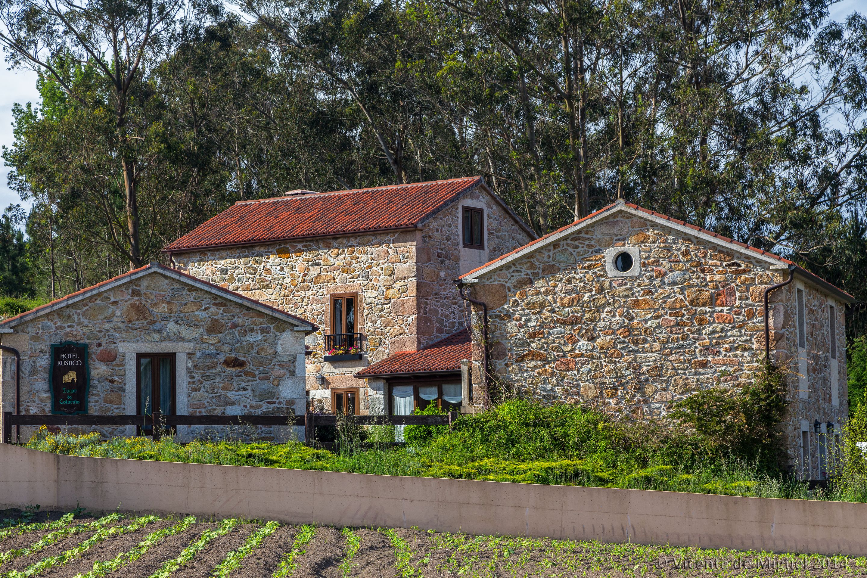 A stone building with a red roof, a sign indicating it is a hotel, and a garden in front.