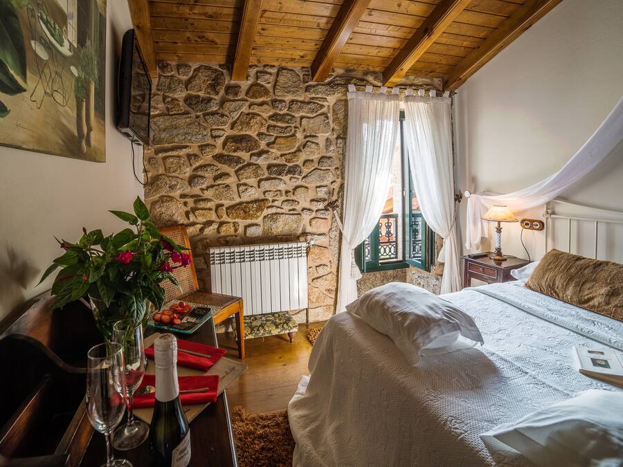 A cozy bedroom with a stone wall, wooden ceiling beams, a bed with white bedding, a bedside table with a lamp, and a window with curtains.
