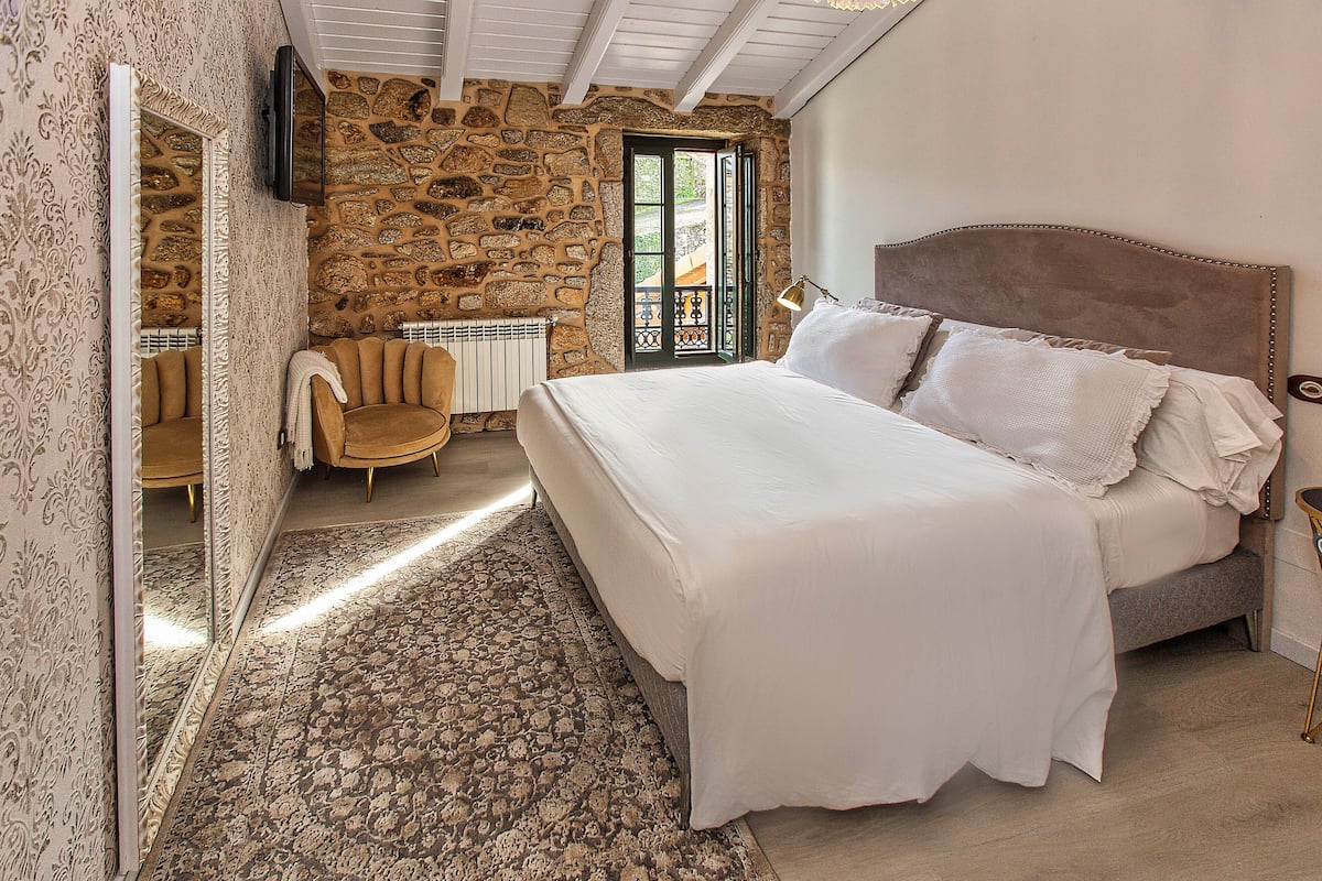 Un dormitorio con pared de piedra, una cama con sábanas blancas, una silla y un espejo.