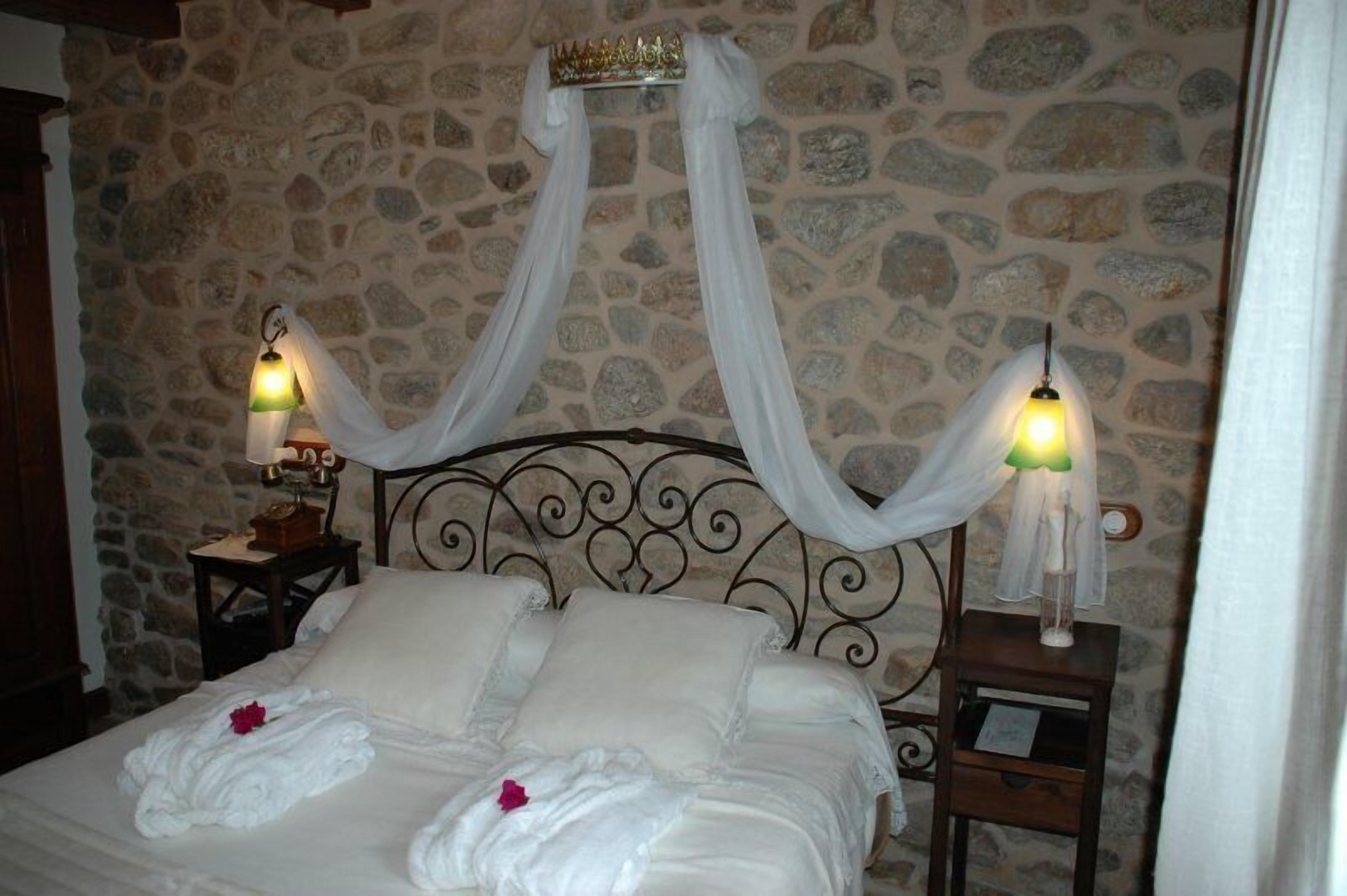 Una cama con ropa blanca y dosel, dos lámparas de noche y un fondo de pared de piedra.