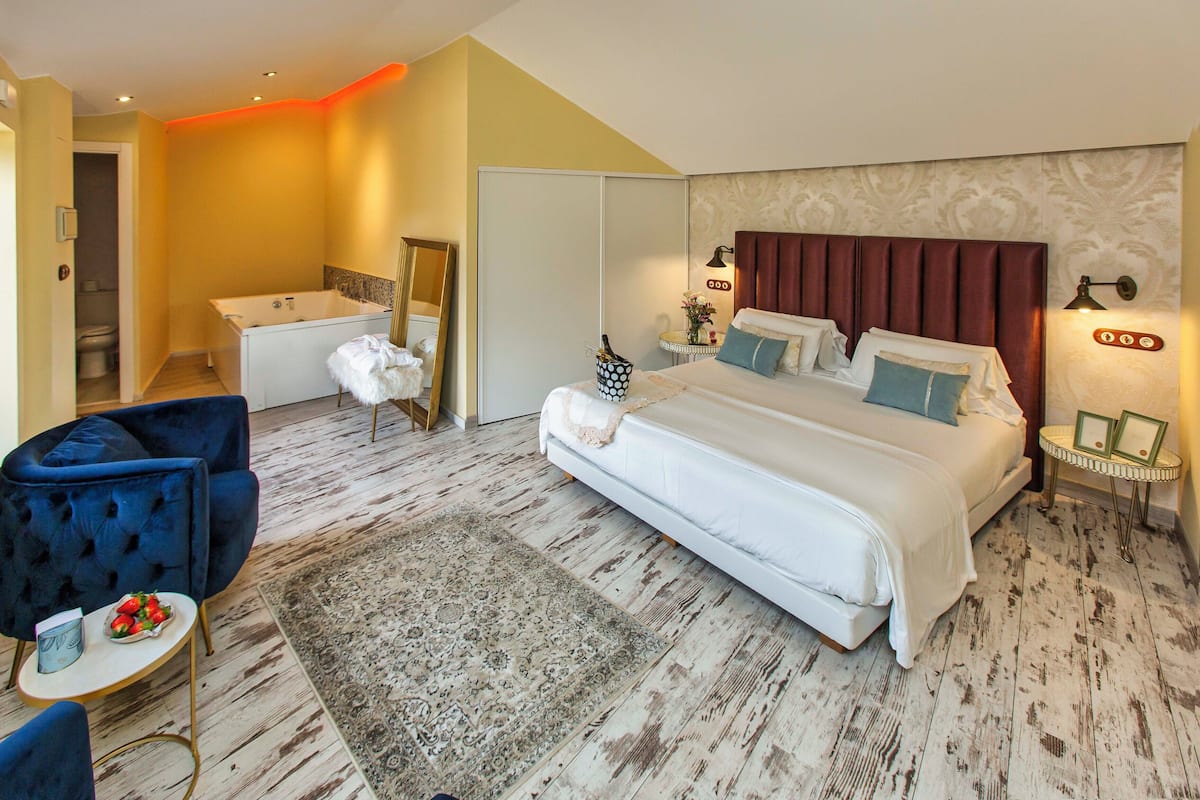 Un dormitorio con una cama grande, una zona de estar con una silla, una mesita con un frutero y una bañera, además de un espejo.