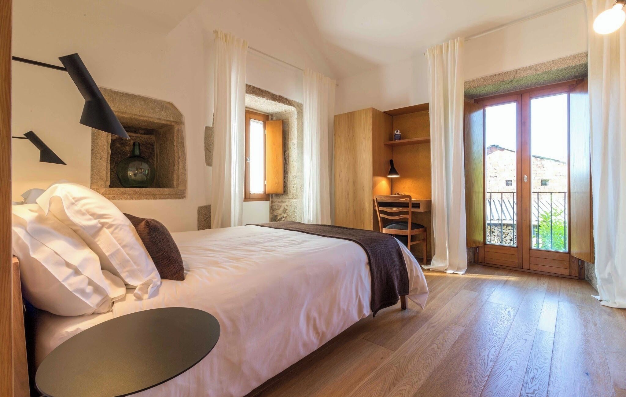 A bedroom with a bed, a desk, a chair, a lamp, and a view of the outside.