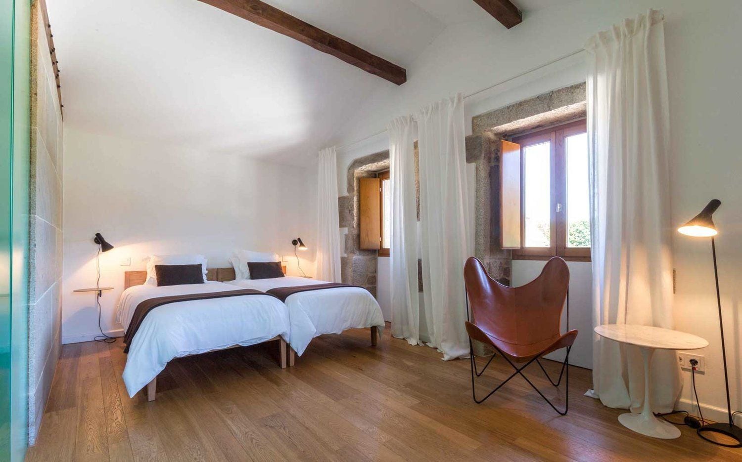 A bedroom with two beds, a chair, a table, and a window with curtains.