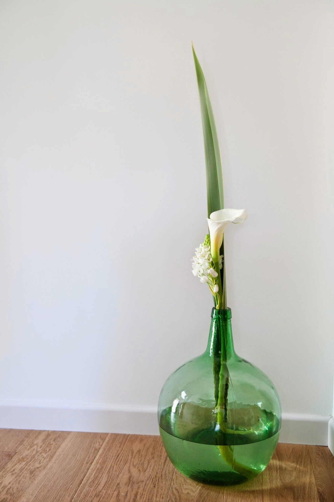Een groene glazen vaas met een witte bloem en groene bladeren op een houten ondergrond.