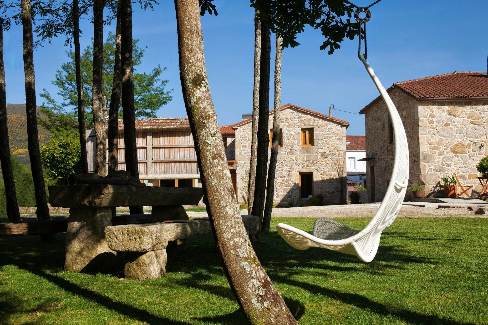 A stone building with a tiled roof, a hammock chair, and a stone bench in a grassy area with trees.