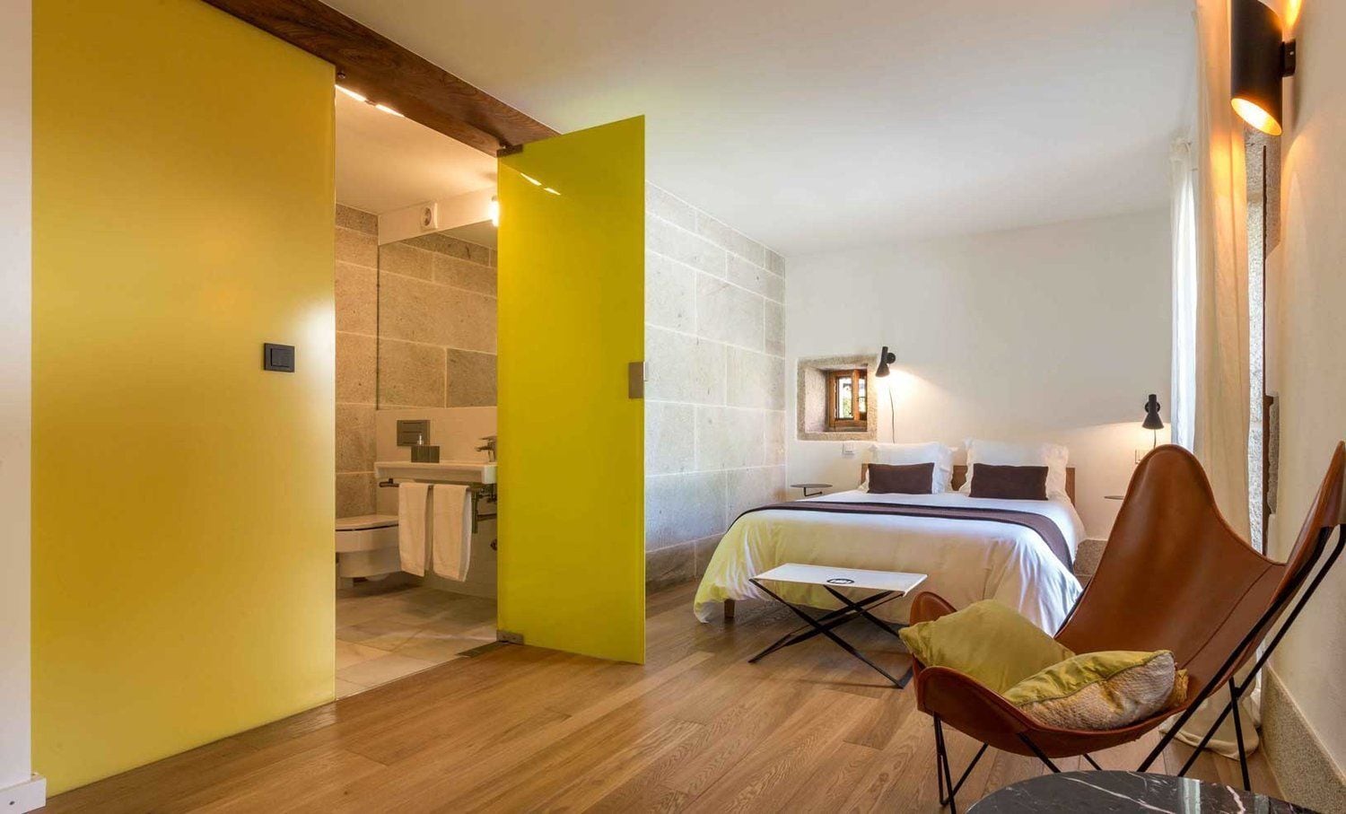 A bedroom with a bed, a chair, a small table, and a bathroom with a toilet and sink.