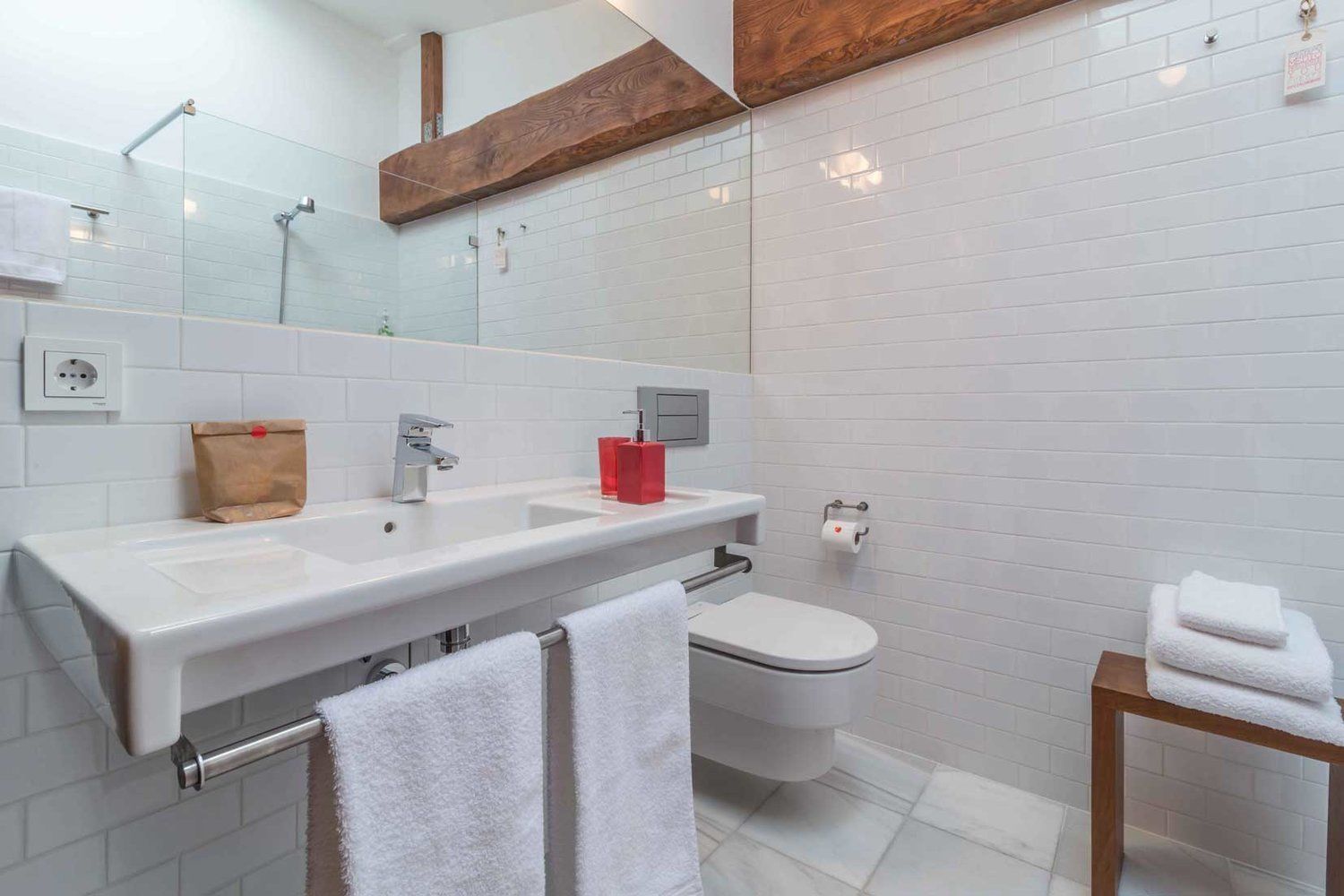 A modern bathroom with a large mirror, two sinks, a toilet, and a wooden shelf above the mirror.