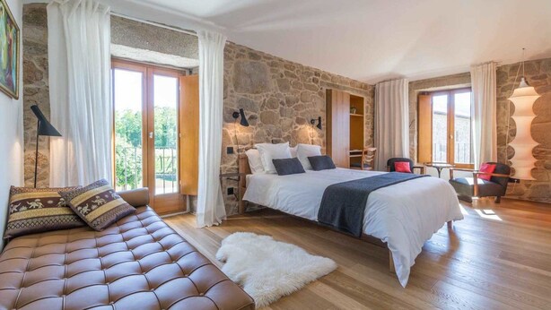 A bedroom with a stone wall, a large bed, a sofa, and a view of the outdoors. - Os Lambráns (Padron)