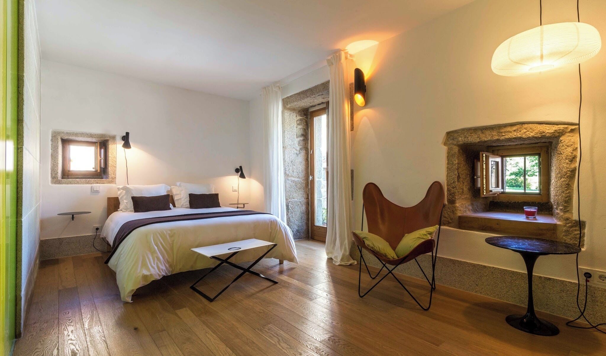 A bedroom with a bed, bedside tables, a chair, a small table, a stone wall, and a window.