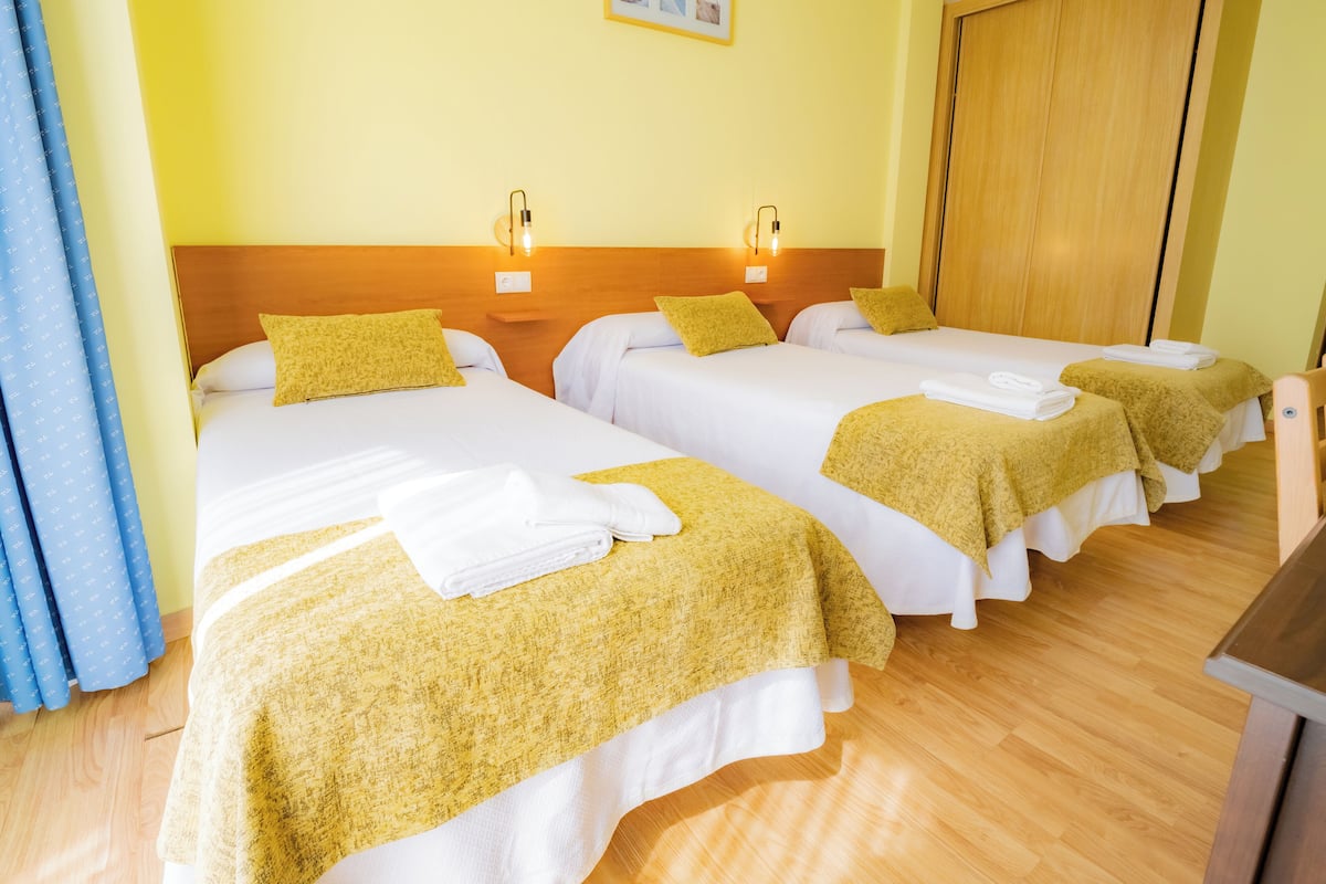 Une chambre d’hôtel avec trois lits simples, chacun doté d’un matelas blanc et d’une couverture jaune. Les lits sont surmontés de têtes de lit en bois, et la chambre comprend une armoire en bois.