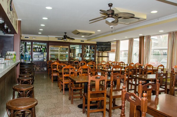 Um restaurante com várias mesas e cadeiras de madeira, ventiladores de teto e um espaço de bar com garrafas e copos.