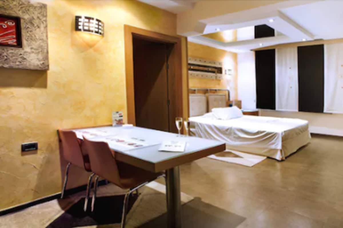 A hotel room with a bed, a desk, and a chair. There is a window with curtains, a wall-mounted light, and a door.