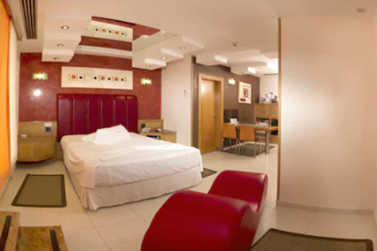 A hotel room with a bed, a desk, and a chair. There is a door leading to another room.