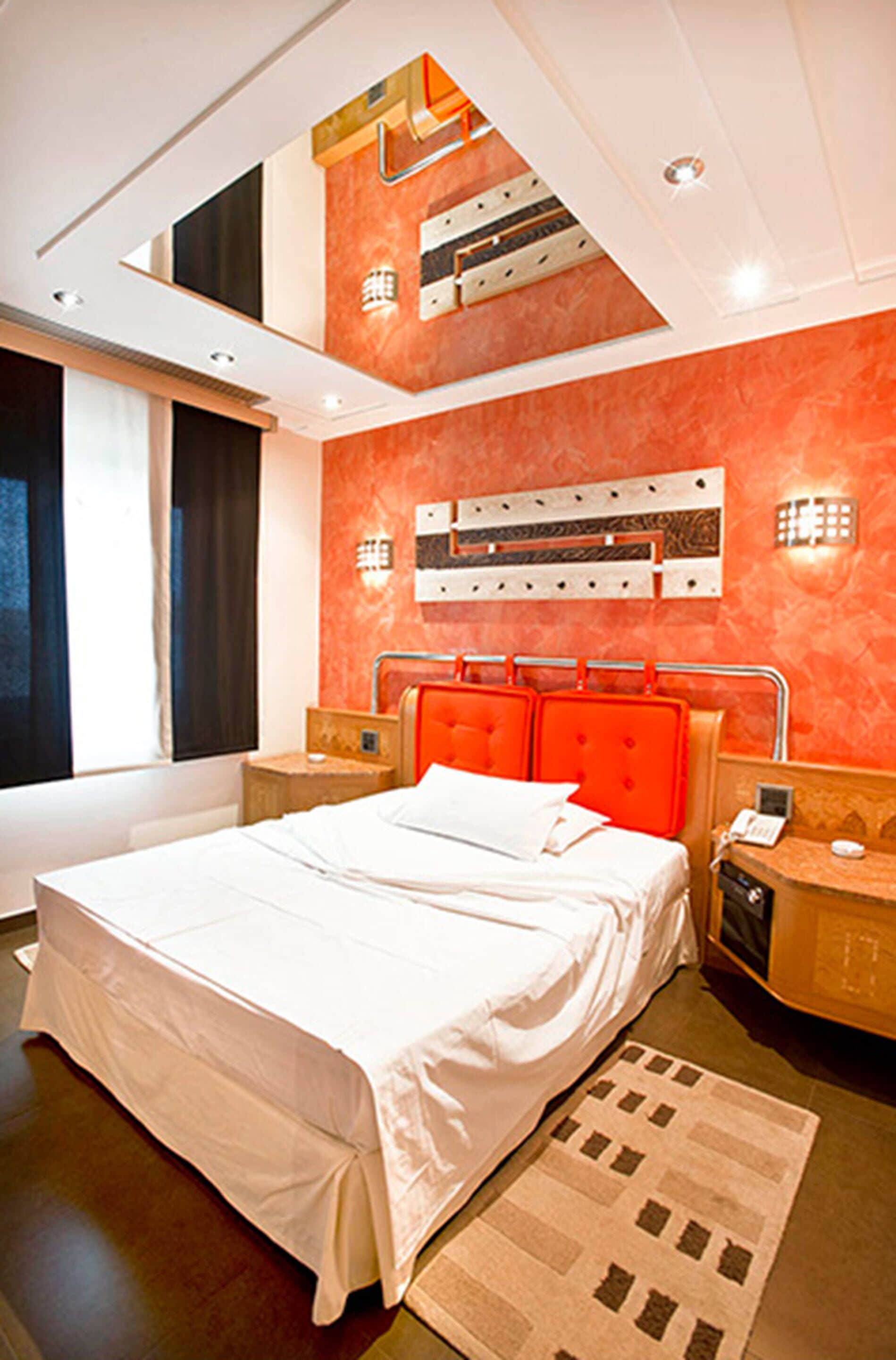 A modern hotel room with a large bed, orange headboard, and a ceiling with recessed lighting.
