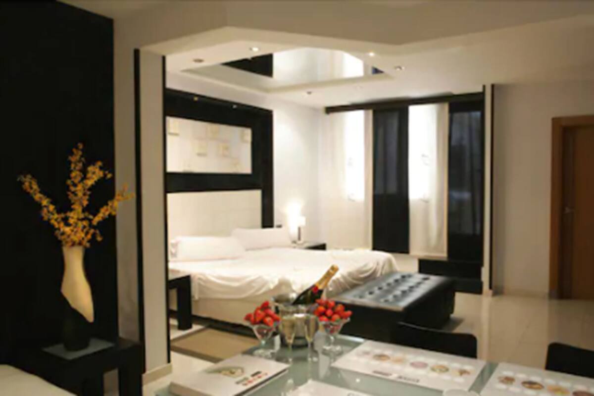 A modern hotel room with a neatly made bed, a bedside table with a champagne bottle, and a small table with fruit and a menu.