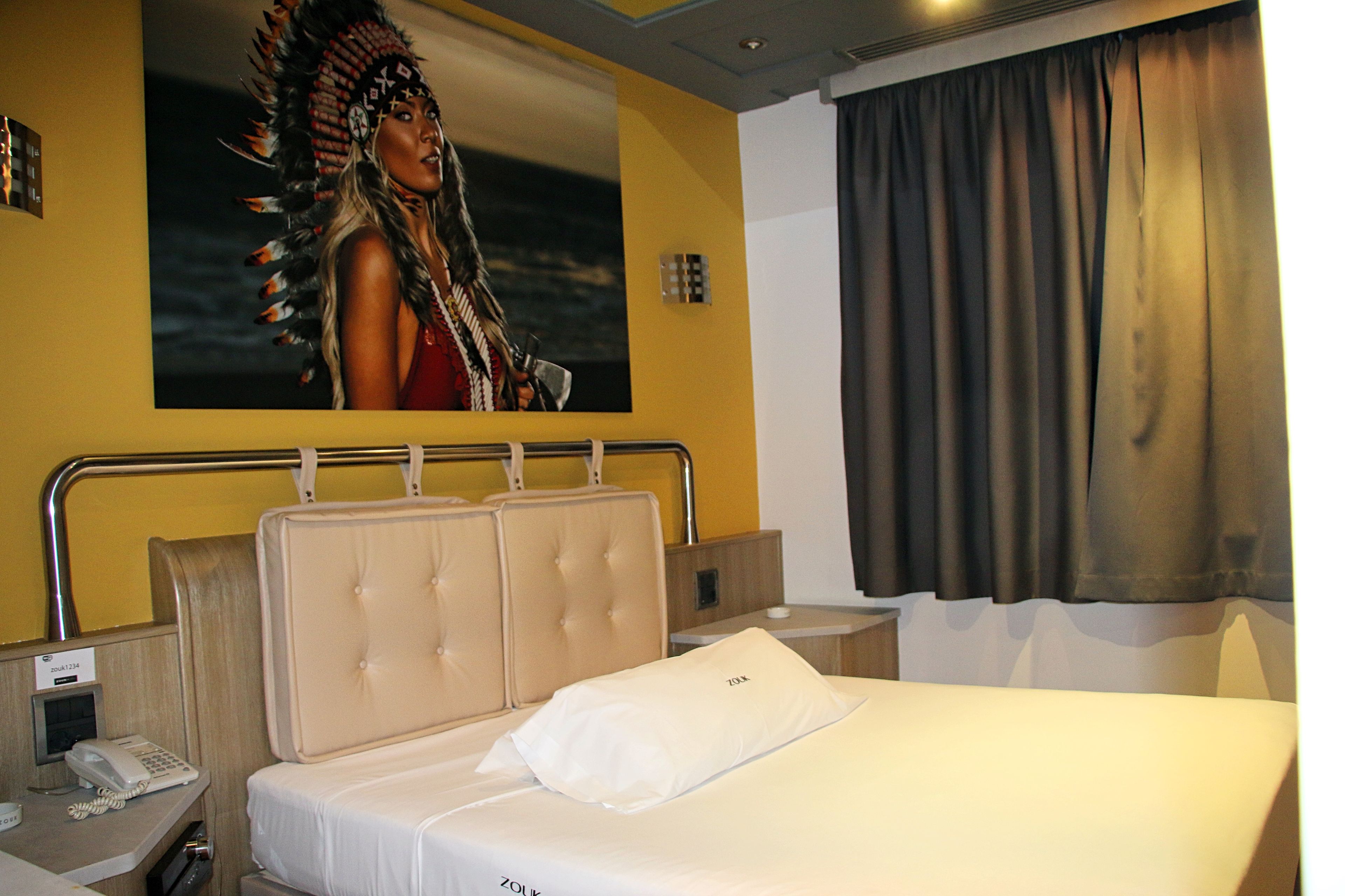Una habitación de hotel con una cama, teléfono, mesita de noche, luz de pared y cortina.