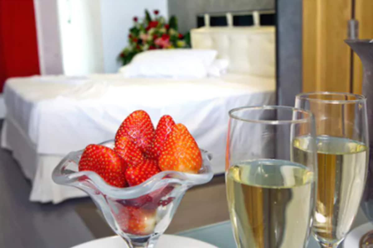 A hotel room with a bed, a glass bowl of strawberries, and two glasses of champagne.