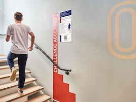 Una persona bajando una escalera con barandilla, un cartel rojo vertical con el texto 'CHALLENGE ACCEPTED' y un código QR en una pared.