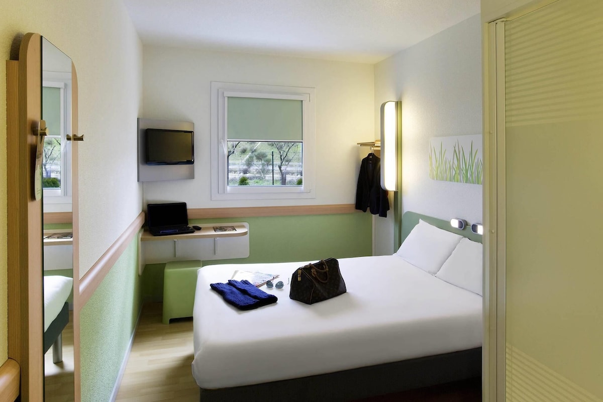 Une chambre d’hôtel avec un lit, un bureau, une télévision, une fenêtre et un miroir.