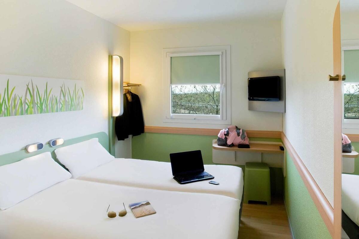 A hotel room with a bed, a laptop, a window, a TV, and a mirror.