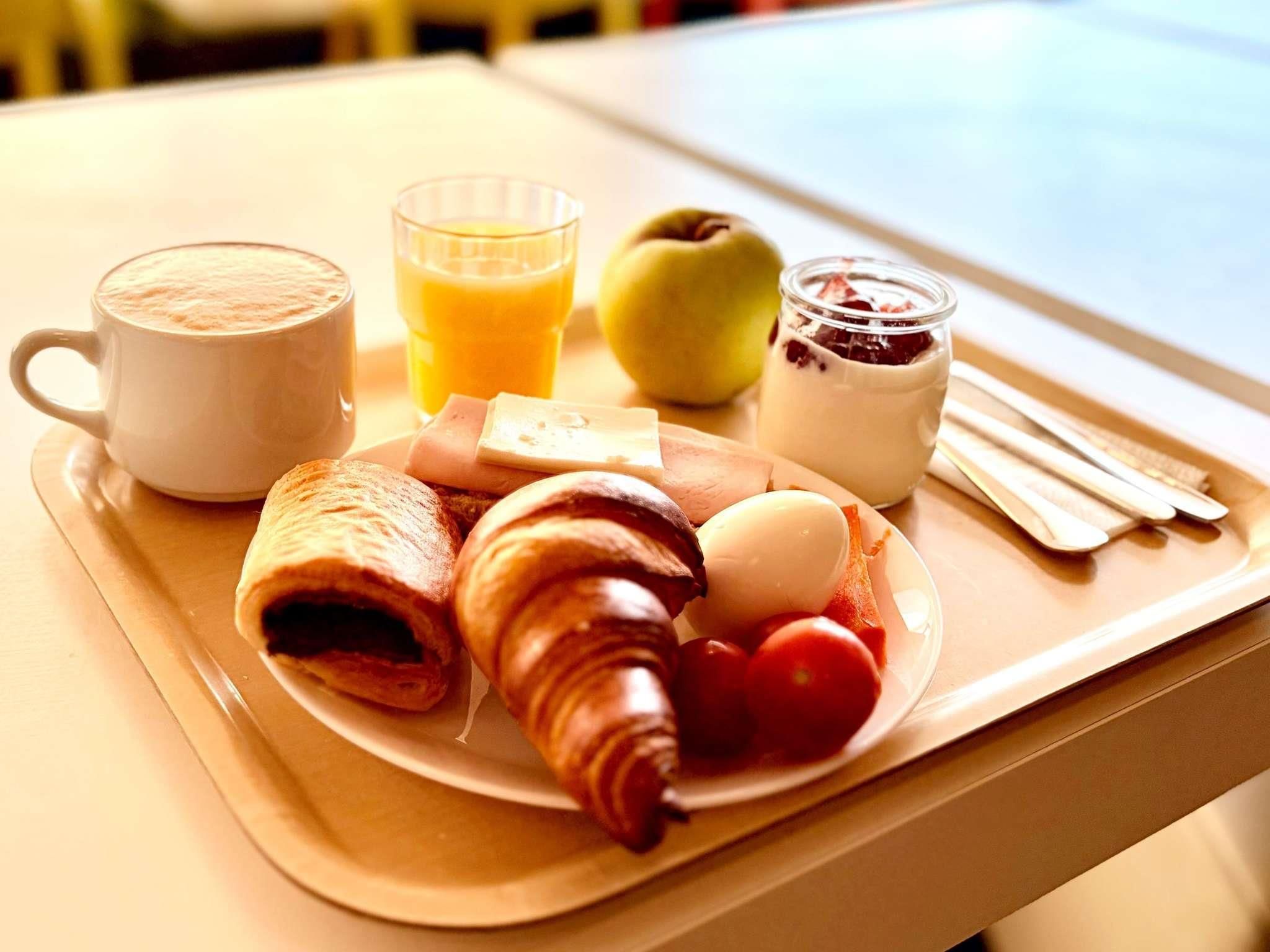Un plateau de petit-déjeuner avec des croissants, un verre de jus d’orange, une tasse de café, une tranche de fromage, une tomate, un œuf dur et un pot de confiture.