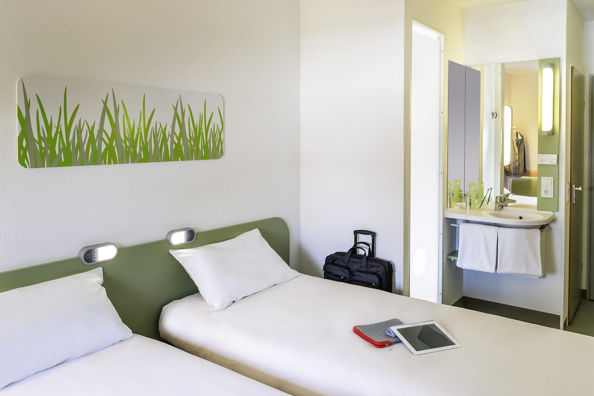 A hotel room with a bed, a bedside table with a lamp, a suitcase, and a bathroom with a mirror and sink.