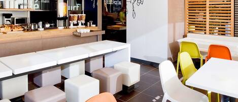 Un café moderno con mesas blancas y sillas coloridas, una barra con diversas opciones de comida y un mural en la pared.