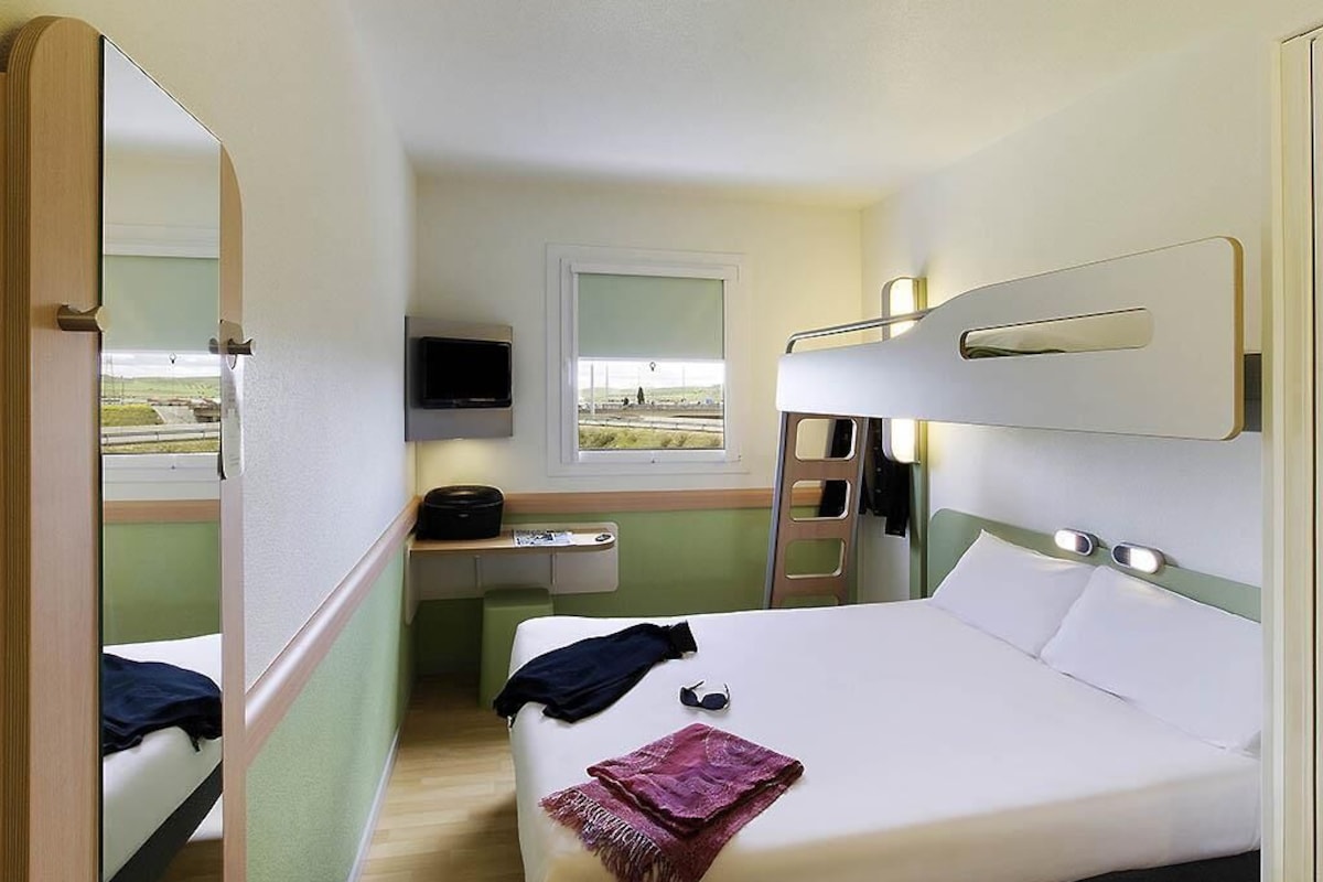 Une chambre d’hôtel compacte comprenant un lit superposé, un petit bureau avec une chaise, une télévision et une fenêtre donnant sur un paysage agréable.