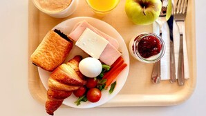 Bandeja de desayuno con cruasanes, jamón, queso, un huevo duro, tomates cherry, lechuga, un vaso de zumo de naranja, una taza de café, una manzana verde y un tarro de mermelada.
