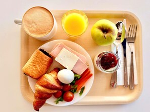 Bandeja de desayuno con cruasanes, jamón, queso, un huevo duro, tomates cherry, lechuga, un vaso de zumo de naranja, una taza de café, una manzana verde y un tarro de mermelada.