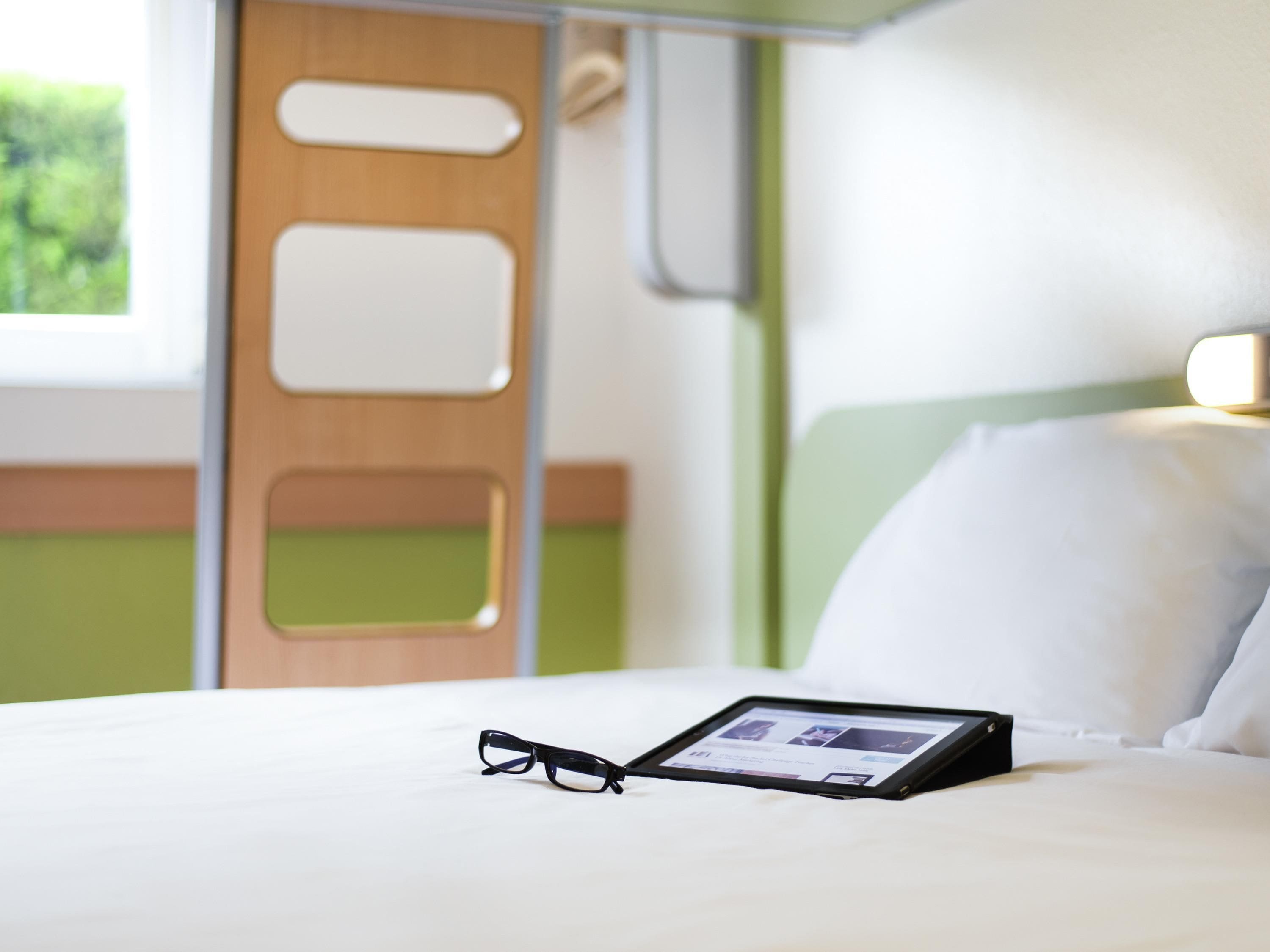 Uma cama com um tablet e óculos, uma parede verde e uma janela com vista.