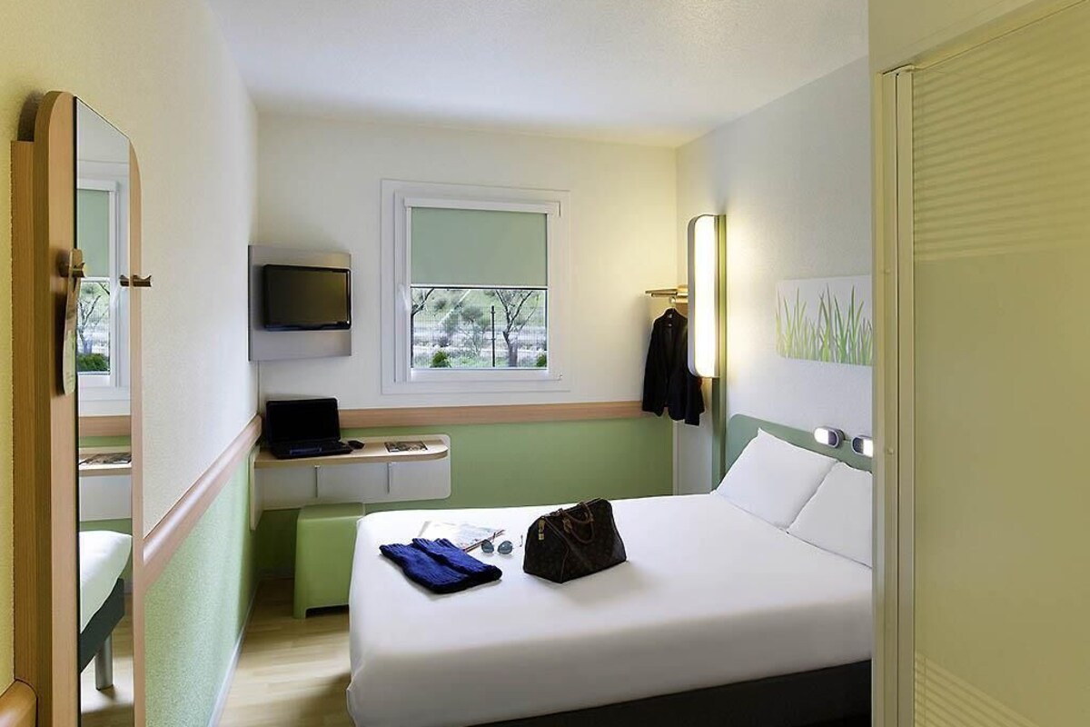 A hotel room with a bed, a desk with a laptop, a TV, and a window with a view of trees.
