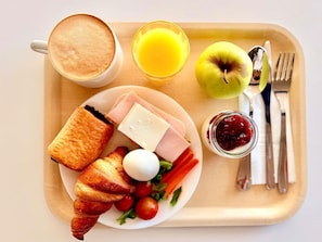 Bandeja de desayuno con cruasanes, jamón, queso, un huevo duro, tomates cherry, lechuga, un vaso de zumo de naranja, una taza de café, una manzana verde y un tarro de mermelada.