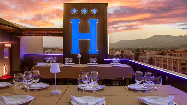 Un espacio de comedor en la azotea con mesas preparadas para una comida, un gran letrero iluminado con la letra 'H' y vistas de la ciudad al atardecer.