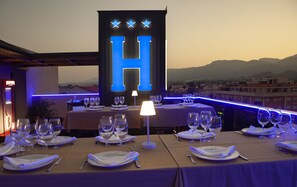 A restaurant with tables set for dining, a large illuminated 'H' sign, and a view of the city and mountains in the background. - Hotel Julián (Alhama de Murcia)