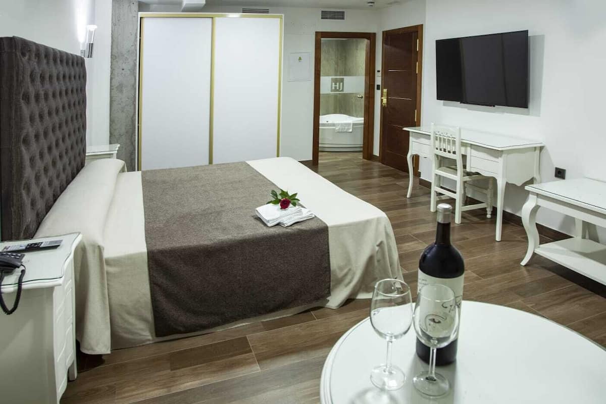 Habitación de hotel con cama, mesita de noche, escritorio, silla, televisor y una botella de vino sobre una mesa.