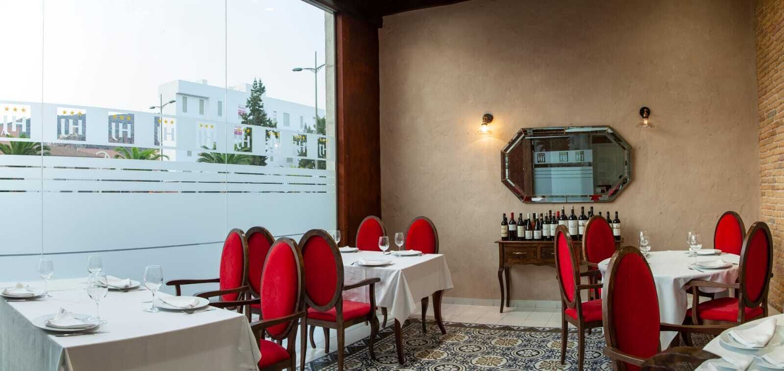 A restaurant with red upholstered chairs and white tablecloths, a brick wall, and a large window overlooking a cityscape.