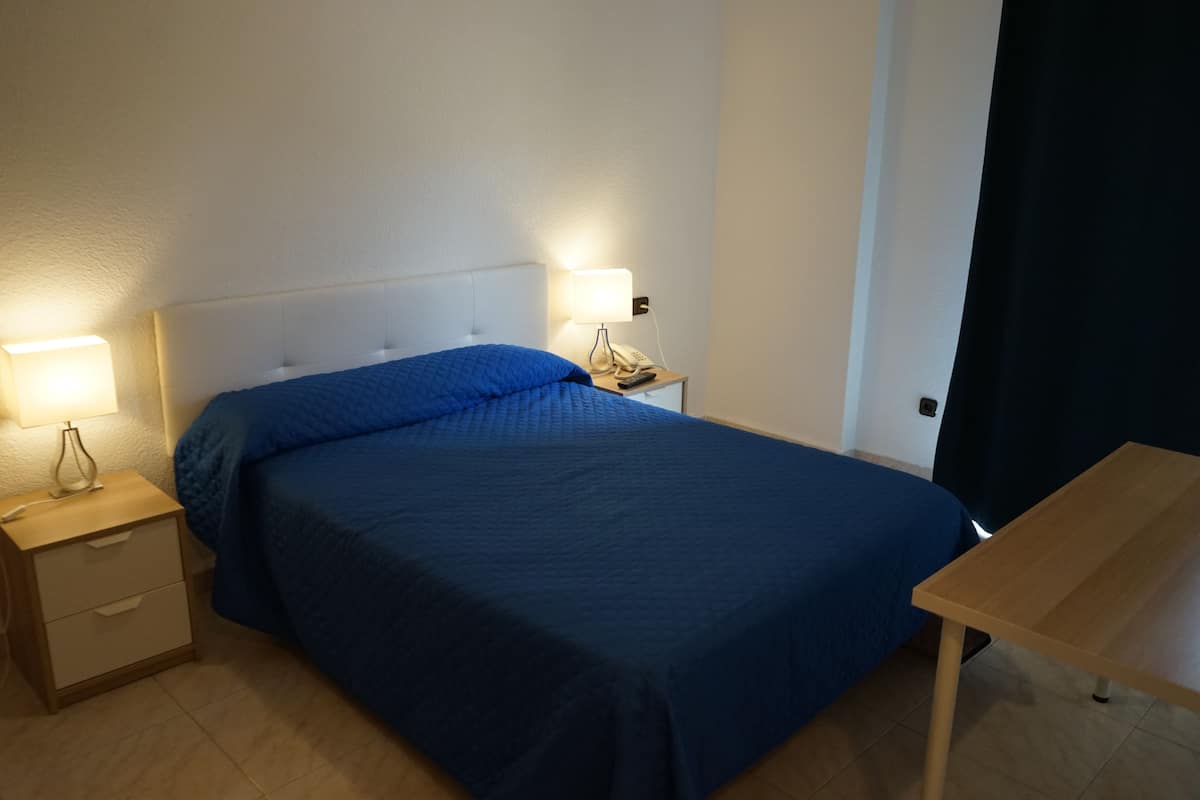 A neatly made bed with a blue bedspread, a bedside table with a lamp, and a small wooden table.