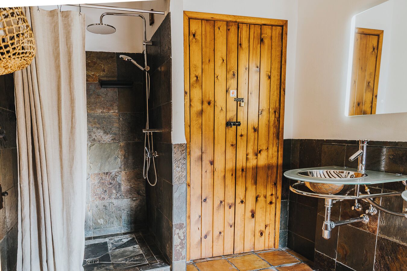 Un baño con ducha de piedra, puerta de madera y lavamanos de vidrio con cuenco de madera.