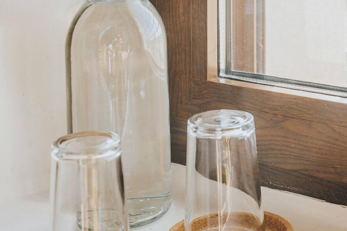 Una botella de vidrio transparente y dos vasos sobre un posavasos de madera junto a una ventana.
