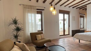 Amplia sala de estar con un sofá, un sillón y una mesita. Se aprecia una ventana grande con cortinas, una planta y una cama al fondo.
