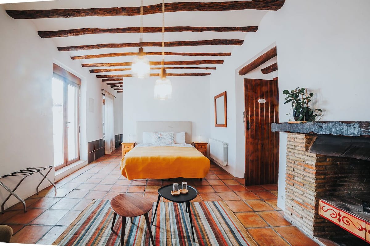 Un dormitorio con cama, una mesita, una silla, chimenea y vistas al exterior.