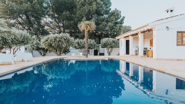 Una piscina con agua azul cristalina, rodeada de sillas reclinables blancas y árboles.
