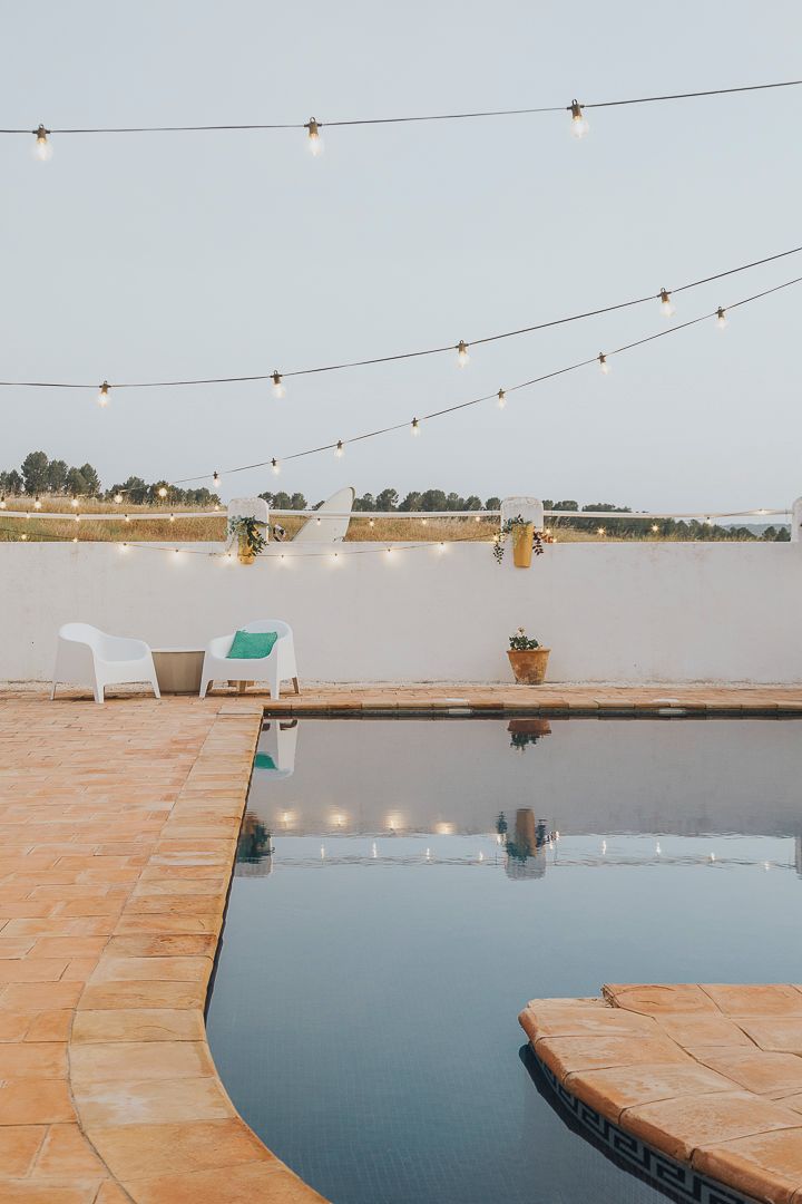 Un área de piscina con luces colgantes, dos sillas blancas y una planta en maceta.
