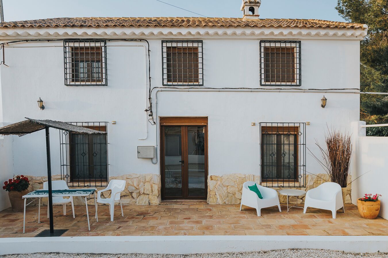 Una casa de dos pisos con techo de tejas, una puerta principal y dos ventanas con rejas. Cuenta con un patio con una mesa y sillas, y una planta en maceta.