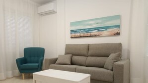 A living room with a grey sofa, a teal armchair, and a white coffee table. A beach scene painting is on the wall. - Vistamar (San Javier)
