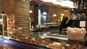 Mostrador de panadería con pasteles y pan, zona de bar con botellas y vasos, y una persona trabajando detrás del mostrador.