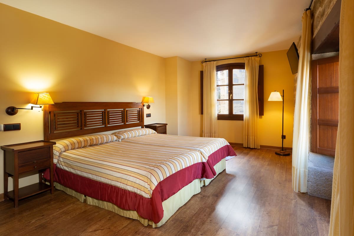 A hotel room with a large bed, wooden headboard, two bedside tables with lamps, a window with curtains, and a wooden floor.