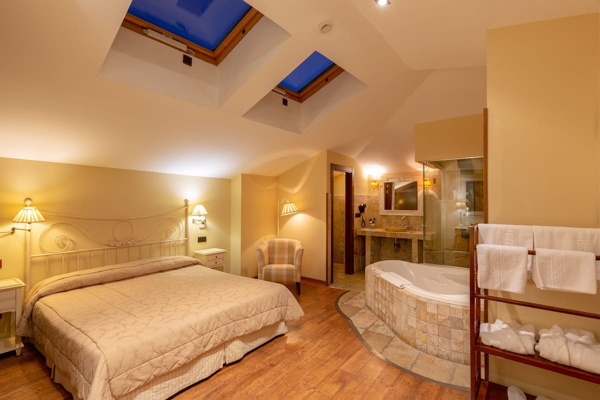 A bedroom with a bed, bedside tables, a chair, a glass-enclosed bathtub, and a towel rack.
